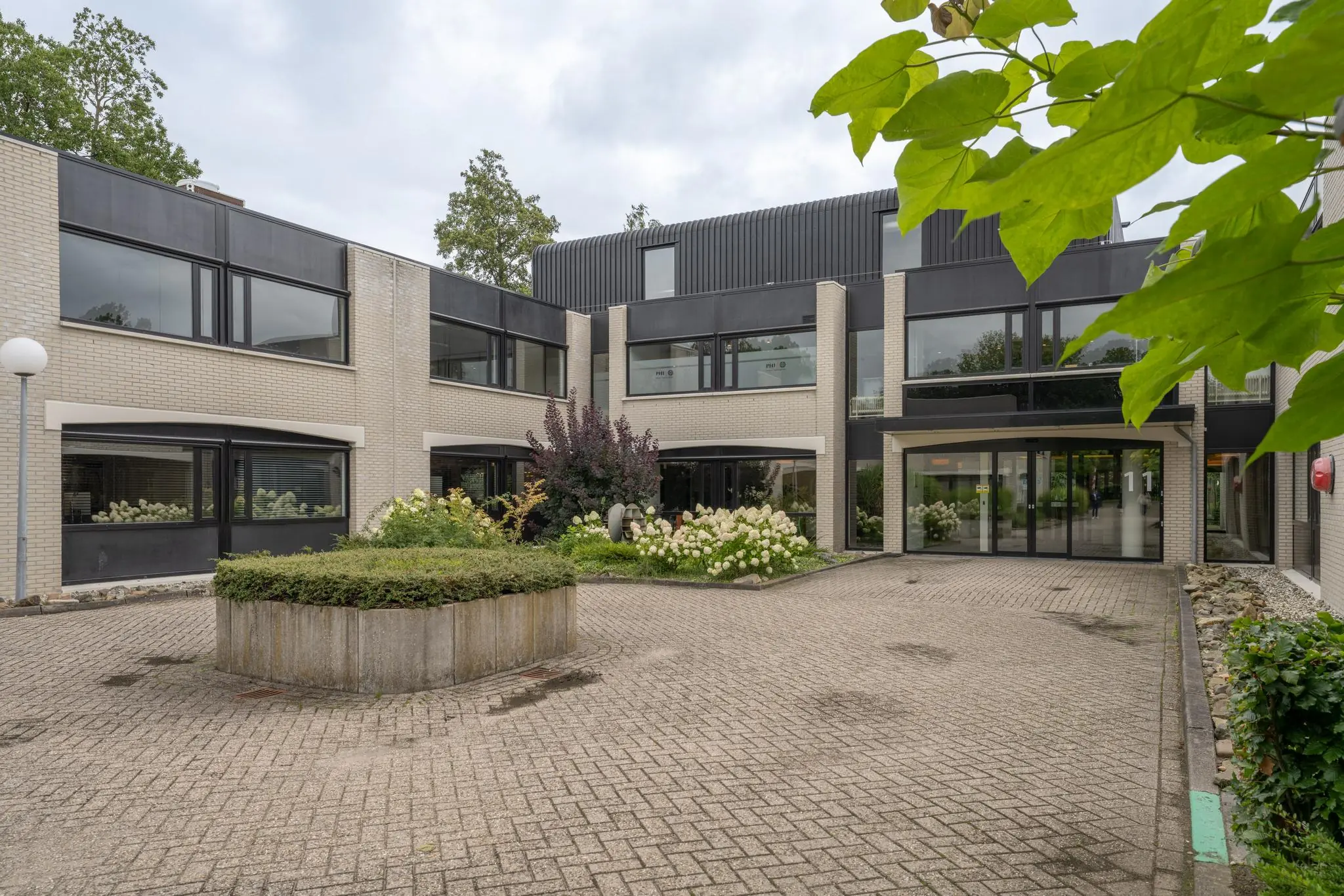 Vooraanzicht van een modern kantoorgebouw aan de Burgemeester Verderlaan 11 met betegelde binnenplaats en bloeiende planten.